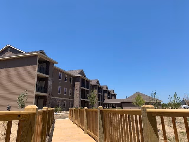 View of a multi-story residential building with balconies under a clear blue sky, seen from a wooden walkway with railings leading towards the building. Small trees and landscaping are visible around the building.