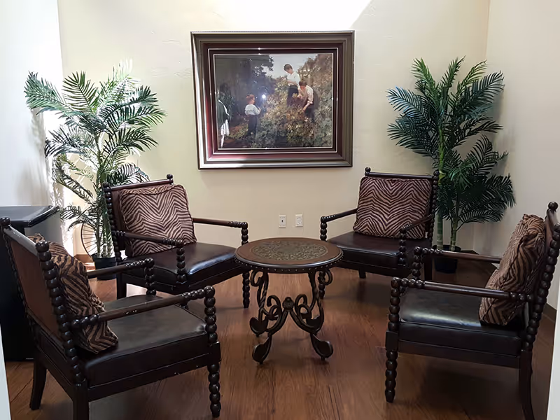 A cozy seating area with four dark wooden chairs featuring patterned cushions arranged around a small round table with ornate legs. Two tall green potted plants are placed against the beige walls, and a framed painting depicting three people in a garden hangs on the wall behind the chairs.