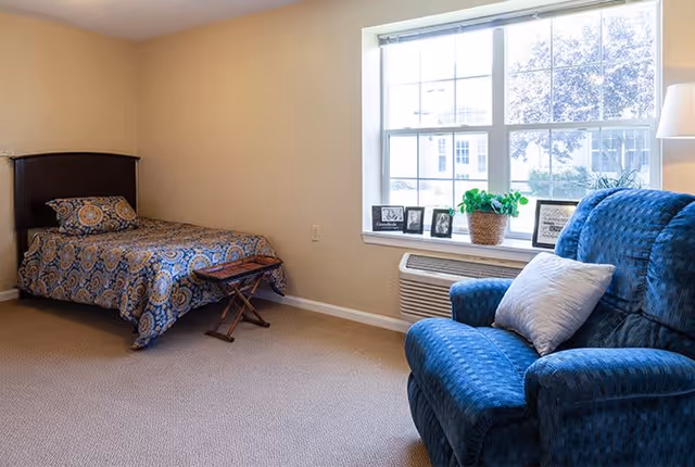 A cozy bedroom in an assisted living facility featuring a single bed with patterned bedding, a small wooden folding table at the foot of the bed, a large blue upholstered armchair with a white pillow, and a window with a windowsill decorated with framed photos and a potted plant.