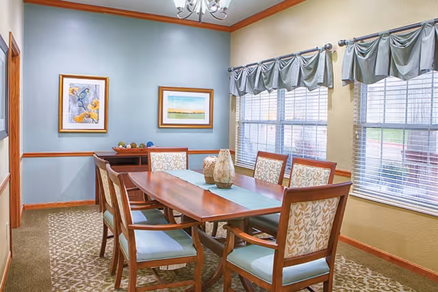 A dining room with a wooden table and six cushioned chairs. The room has light blue and beige walls, two framed paintings on the wall, three large windows with valance curtains, and a chandelier overhead. Decorative vases are placed on the table.