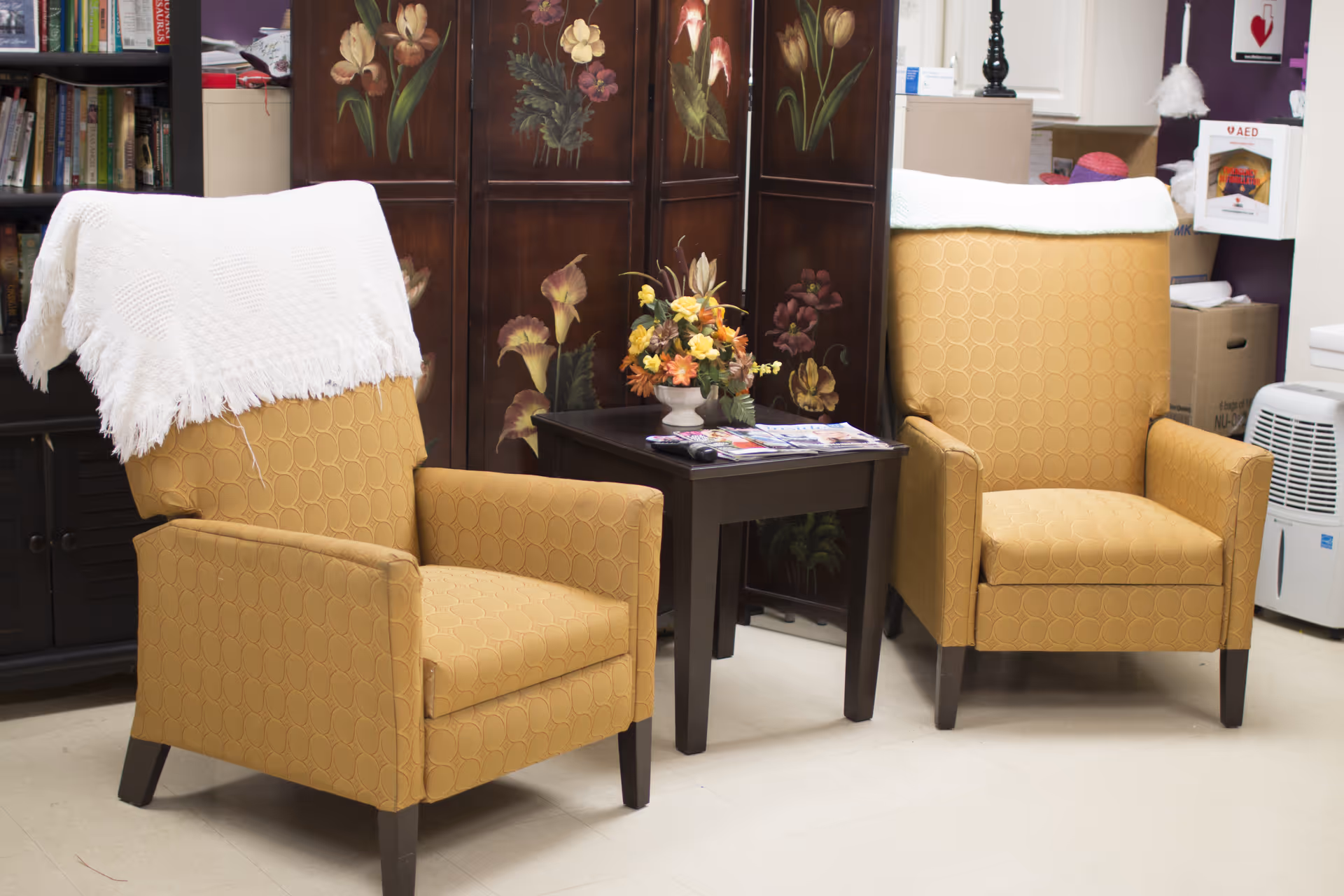 Two mustard yellow armchairs with a white throw draped over one, positioned around a small dark wooden table with a floral arrangement and magazines on top, set against a decorative folding screen with floral paintings and a bookshelf in the background.