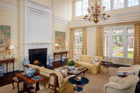 A spacious and elegant living room with high ceilings and large windows allowing natural light to fill the space. The room features a lit fireplace with a white mantel, two beige sofas, two armchairs, and several wooden side tables adorned with blue and white ceramic vases and lamps. The walls are decorated with framed artwork, and a large chandelier hangs from the ceiling. Outside the windows, a garden and walkway are visible.