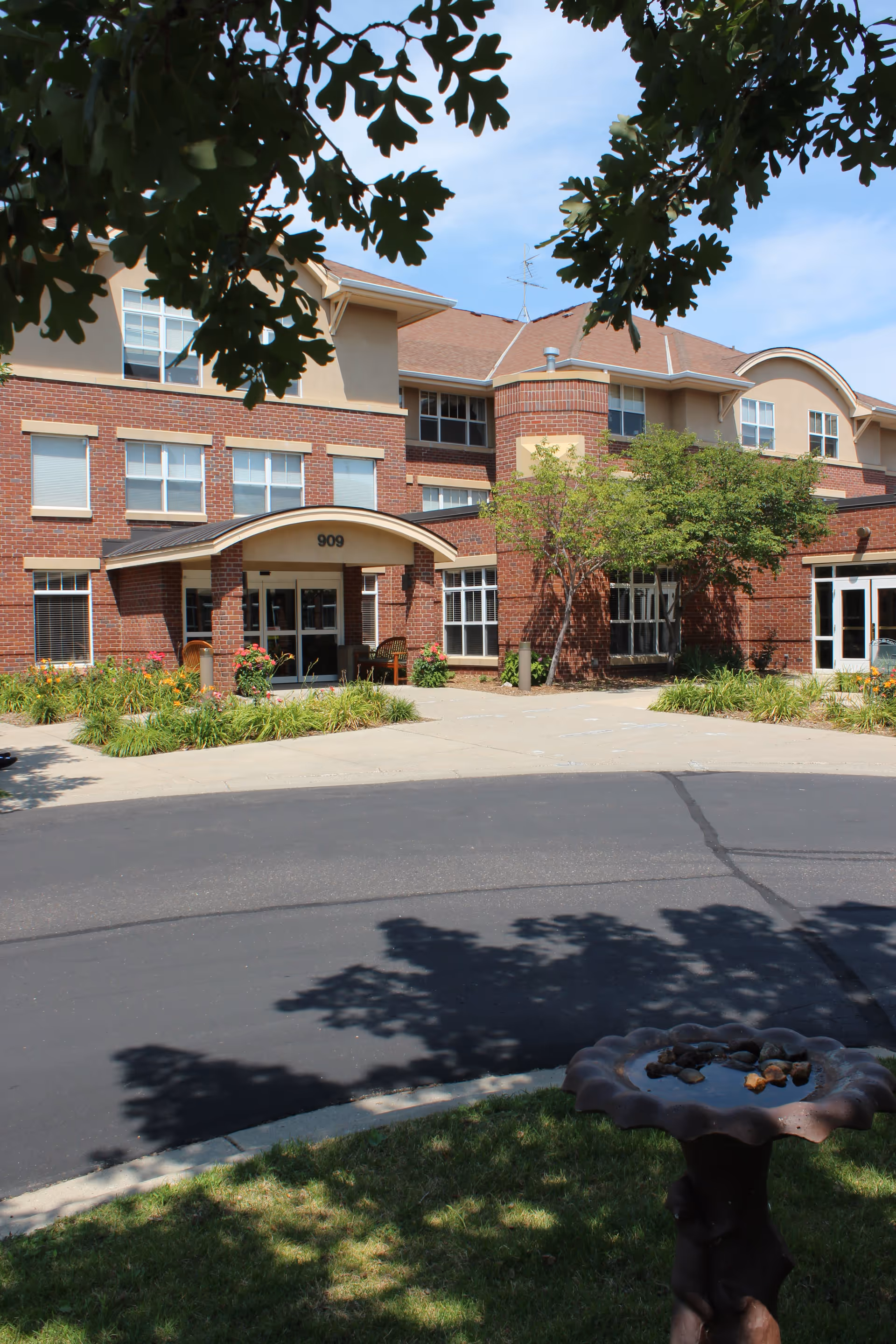 Front exterior view of a multi-story senior living facility named Main Street Lodge with a brick and beige facade, a covered entrance with the number 909 above it, surrounded by landscaped greenery and trees, and a birdbath in the foreground.