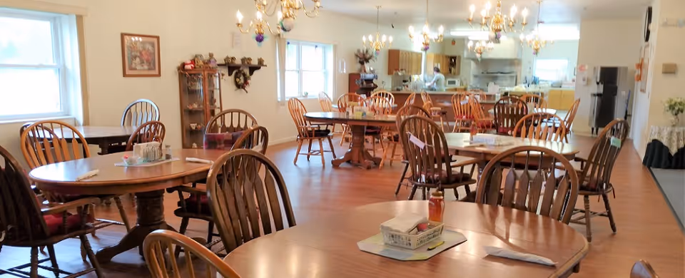 Bright communal dining room with multiple wooden tables and chairs and a kitchen area in the back.