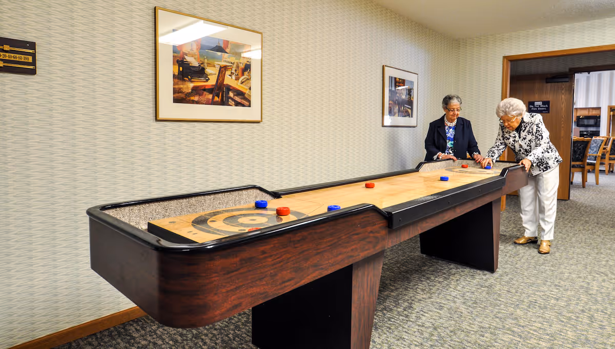 Two elderly women playing shuffleboard on an indoor shuffleboard table in a carpeted room with framed artwork on the walls. One woman is pushing a red puck while the other watches. There is a doorway leading to another room with chairs and tables in the background.
