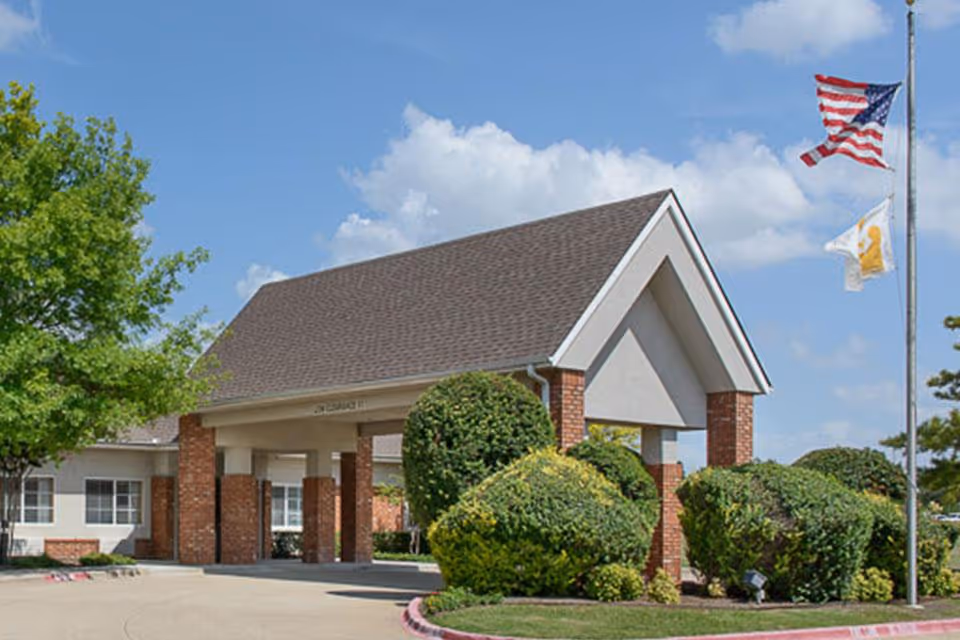 Front entrance of a care facility with a covered portico, trimmed shrubs, and flags flying on a flagpole.
