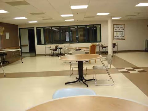 Empty dining room in senior facility with round tables, chairs, and large windows.