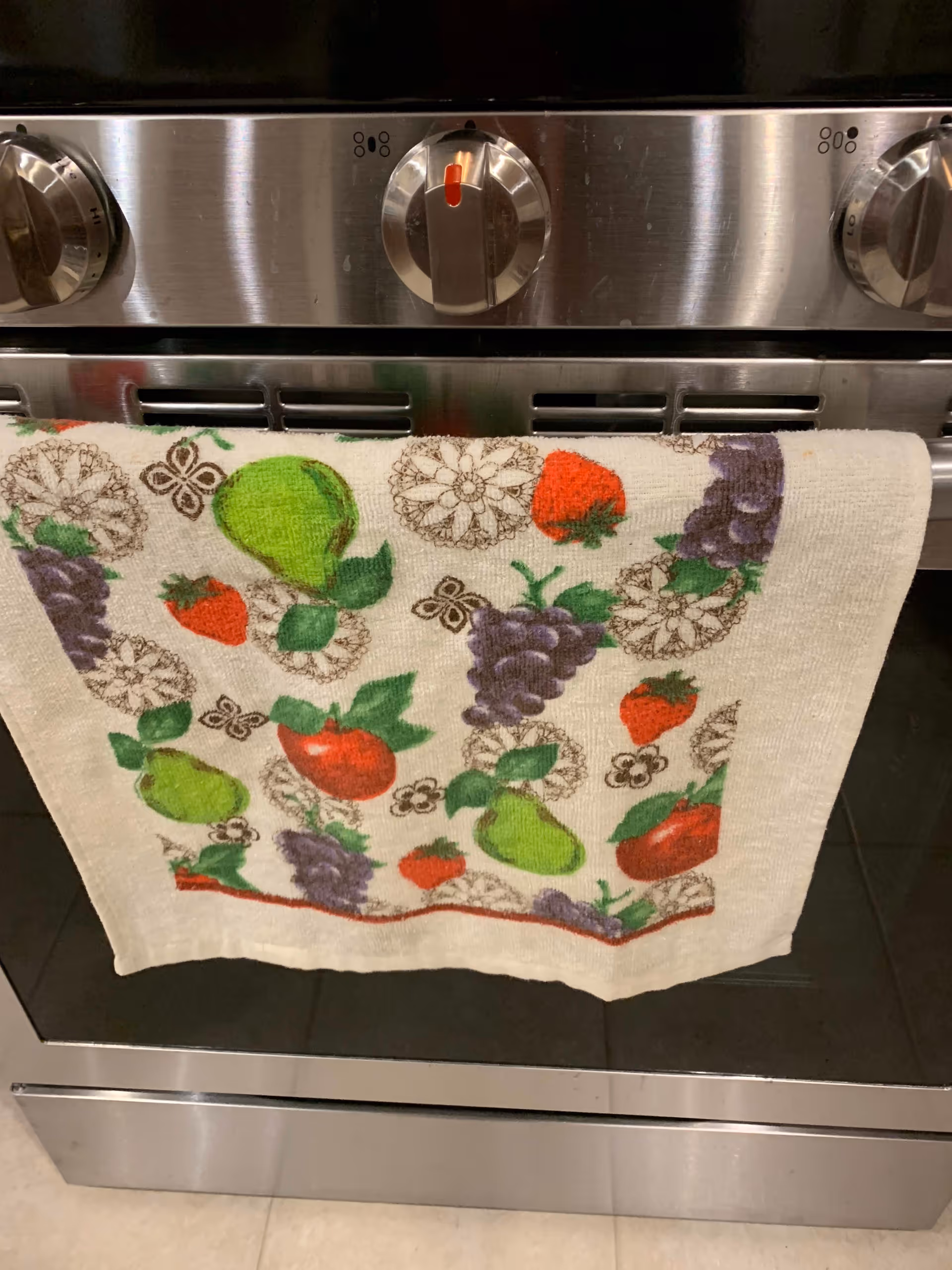 Close-up view of a stainless steel oven with three control knobs. A white kitchen towel decorated with colorful fruit patterns including grapes, strawberries, pears, and apples hangs from the oven handle.