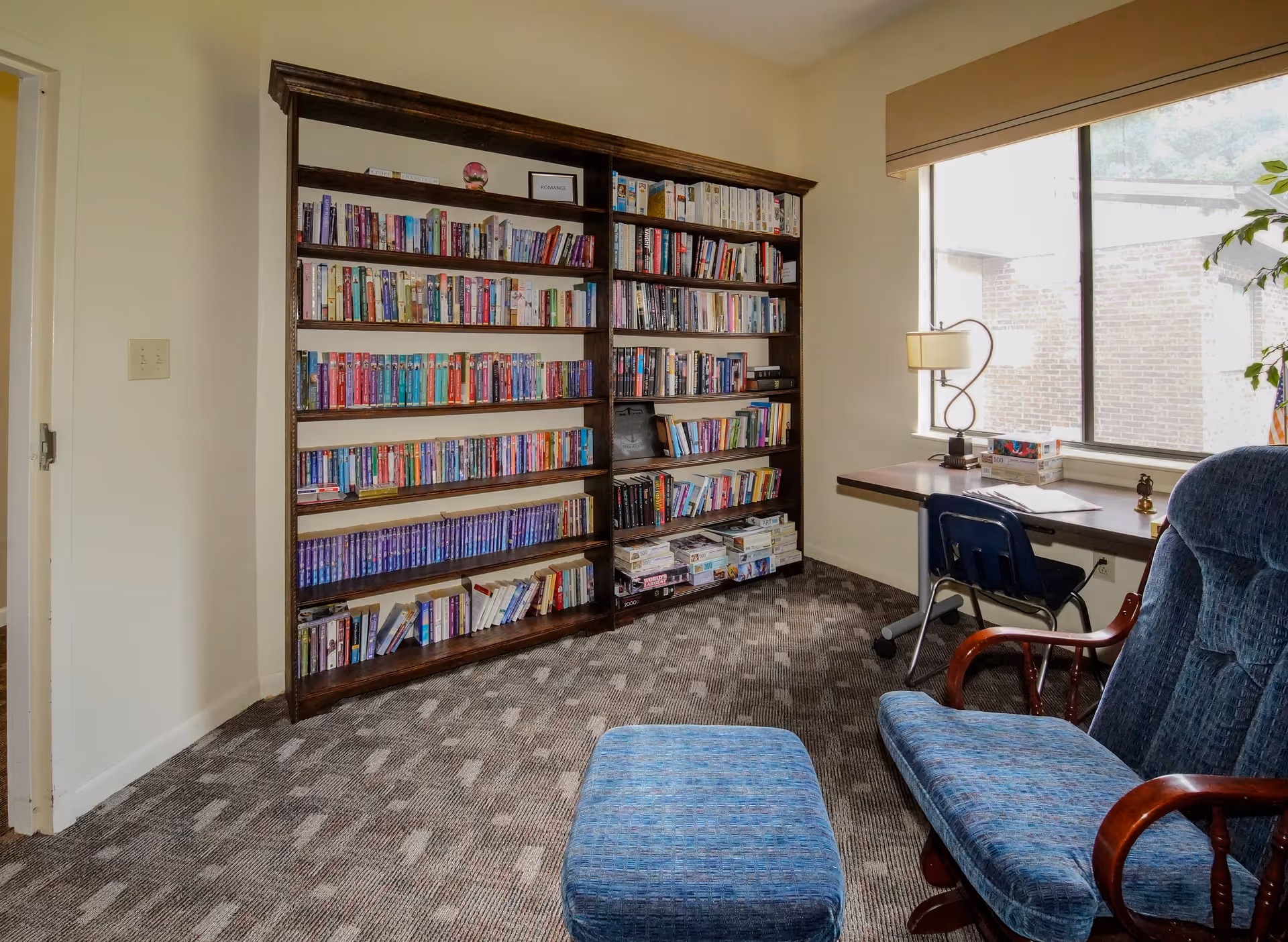 A cozy reading area with a large bookshelf, a desk by a window, and an upholstered chair with an ottoman.