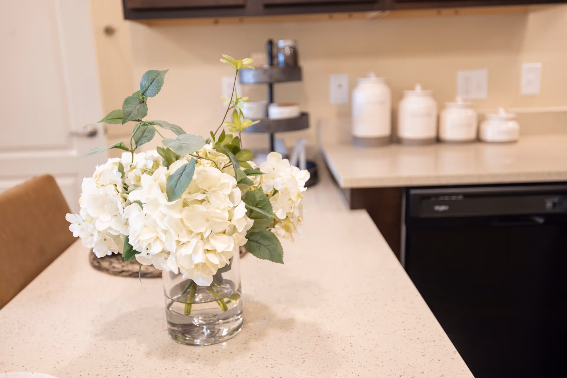 A close-up view of a kitchen countertop with a clear glass vase holding white hydrangea flowers and green leaves. In the background, there are white ceramic canisters and a tiered black stand on the countertop, along with dark wood cabinets and a black dishwasher.