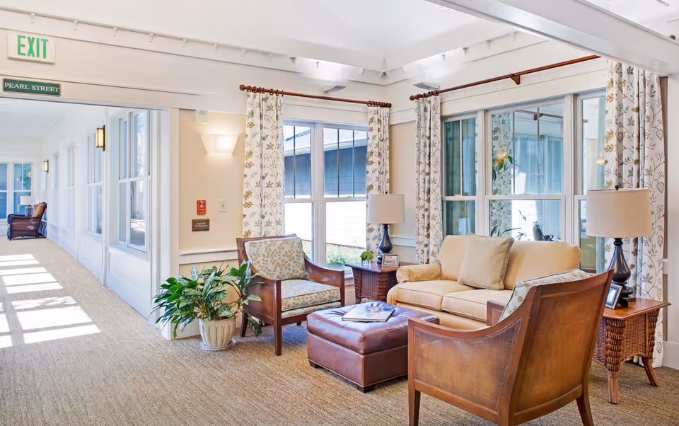 Bright, airy common lounge with a sofa, armchairs, an ottoman, side tables and lamps beside large windows and a sunlit corridor.