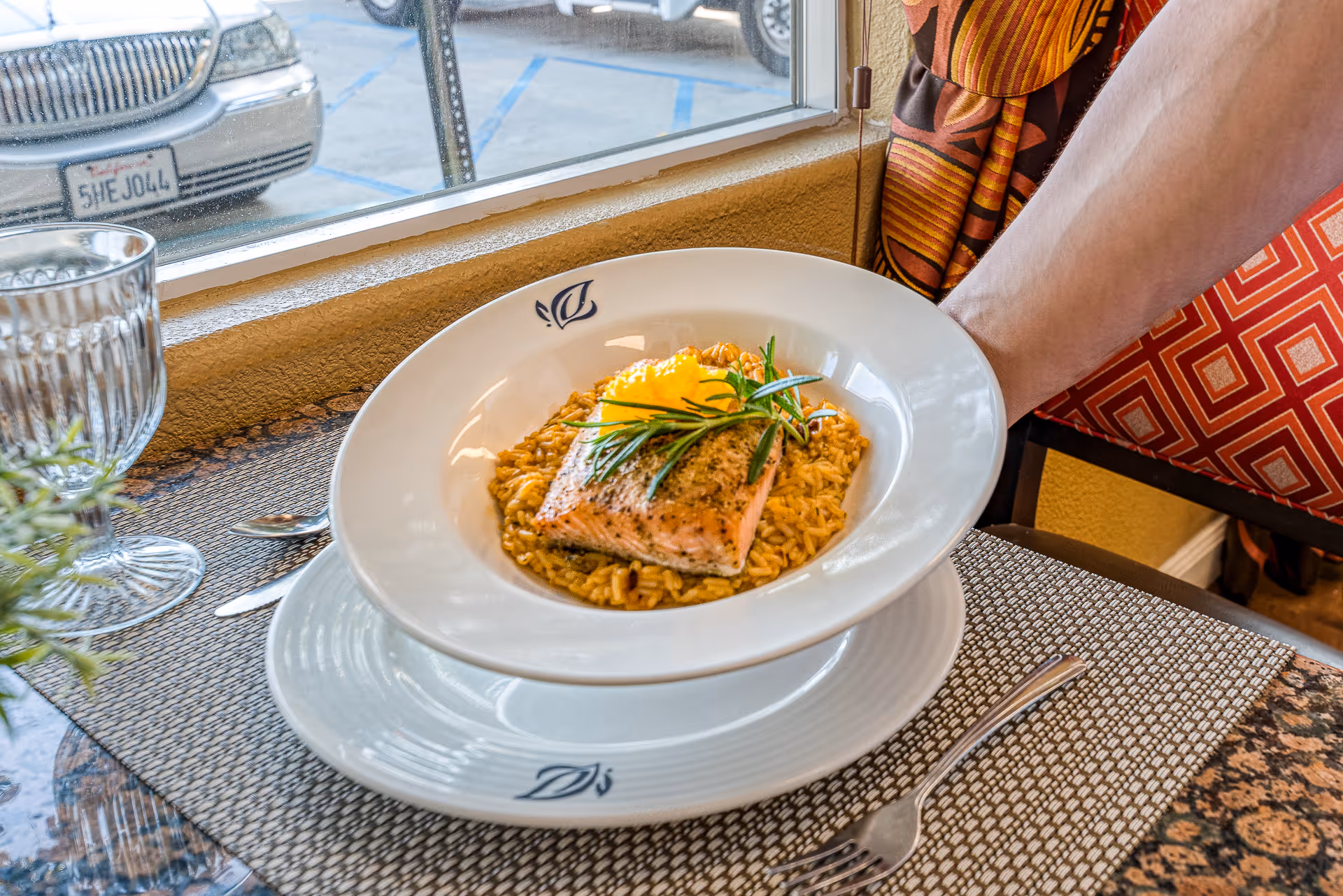 A plate of cooked salmon garnished with a sprig of rosemary and a lemon slice, served on a bed of rice. The plate is being held by a person wearing a patterned shirt, next to a window with a view of parked cars outside. A glass and silverware are placed on the table.