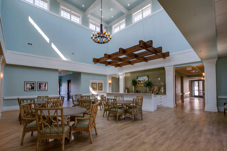 Bright communal dining room with round wooden tables and chairs under a high ceiling, chandelier and decorative wooden beams.