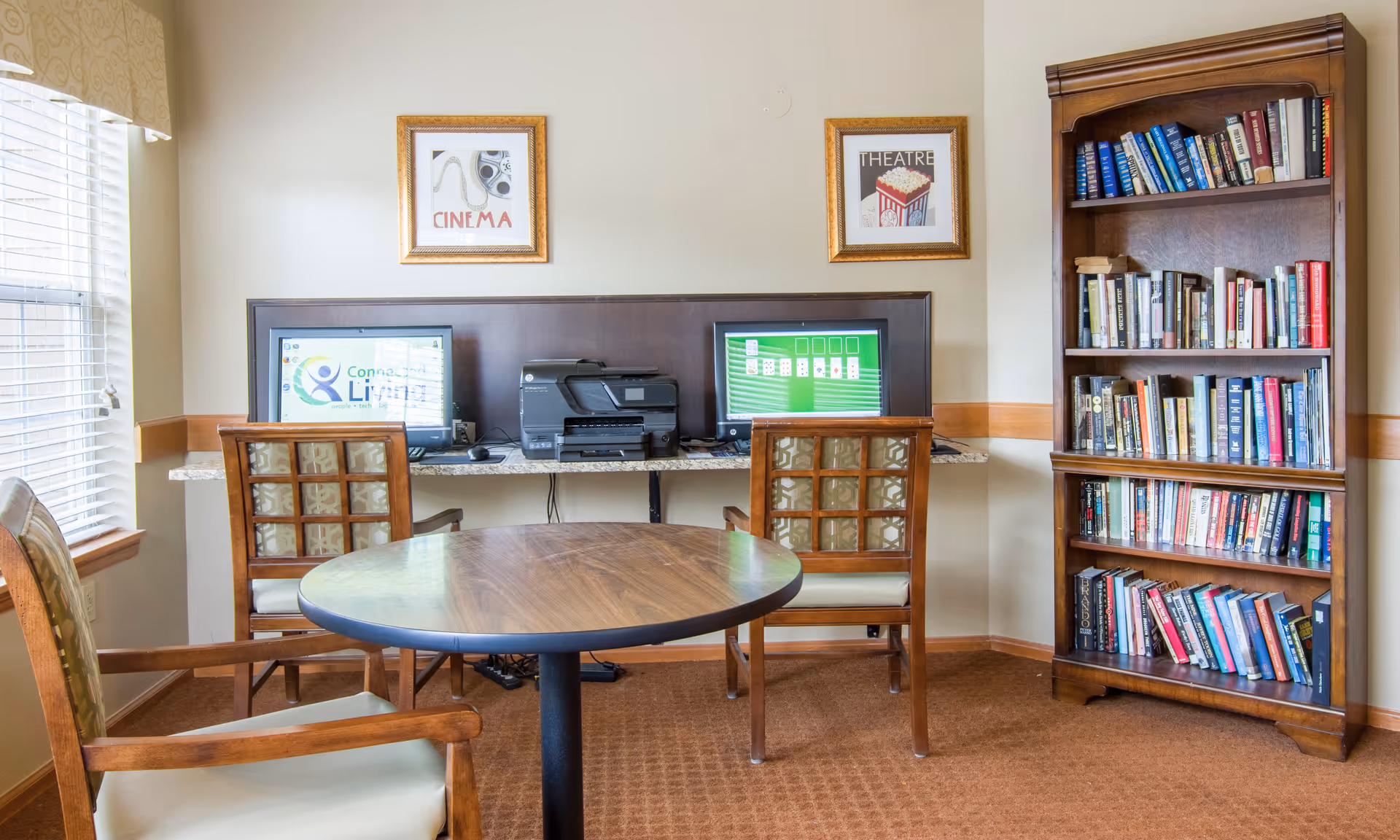A cozy room with a round wooden table and three wooden chairs with cushioned seats. Against the wall, there is a granite countertop with two computer monitors and a printer. Above the countertop, two framed pictures hang on the wall, one labeled 'CINEMA' and the other 'THEATRE'. To the right, there is a tall wooden bookshelf filled with books. A window with blinds is on the left side of the room, allowing natural light to enter.