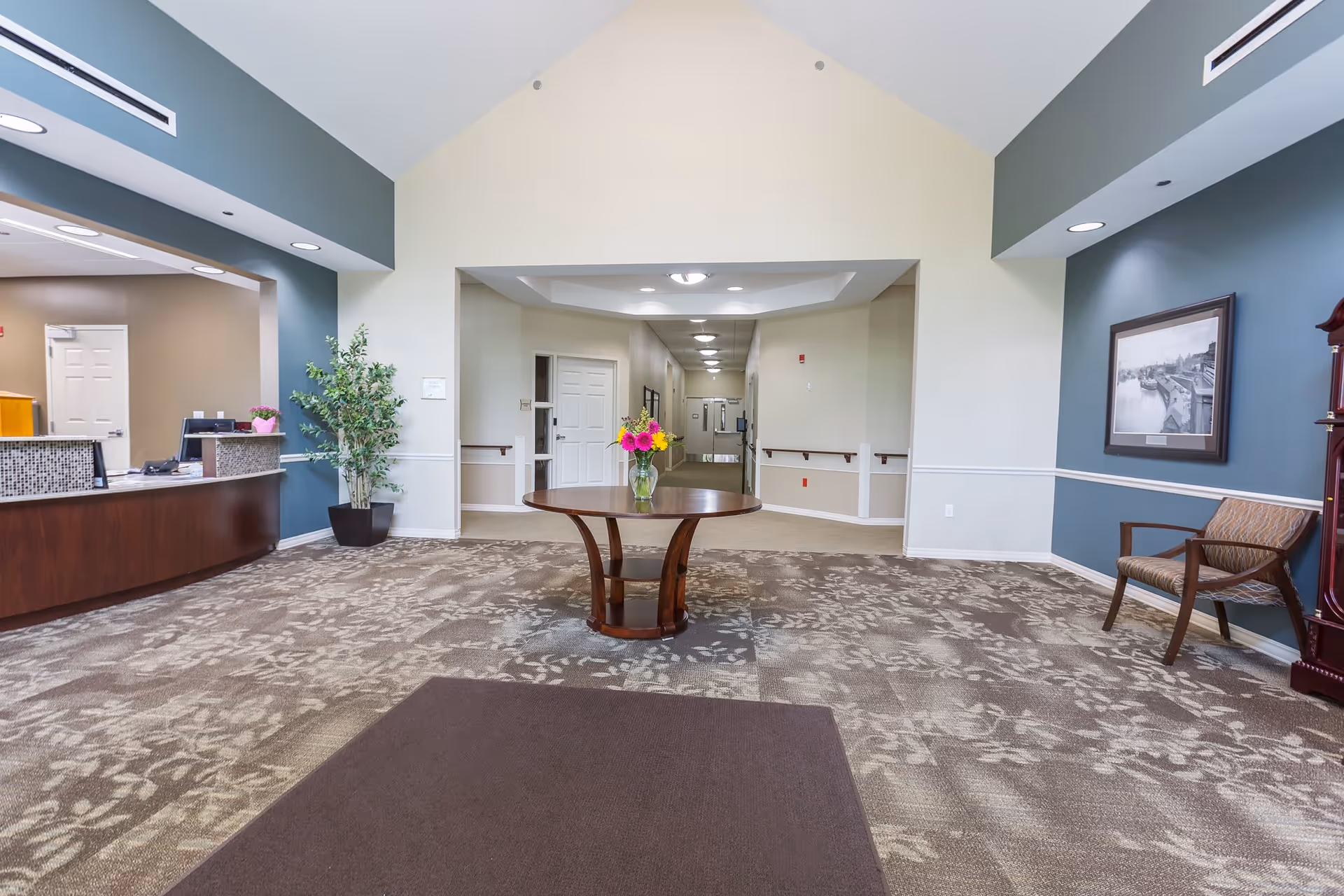 Interior view of a senior living facility lobby with a high vaulted ceiling, a round wooden table with a vase of colorful flowers in the center, a reception desk on the left, a potted plant, a framed black and white photo on the right wall, and a hallway leading to other rooms.