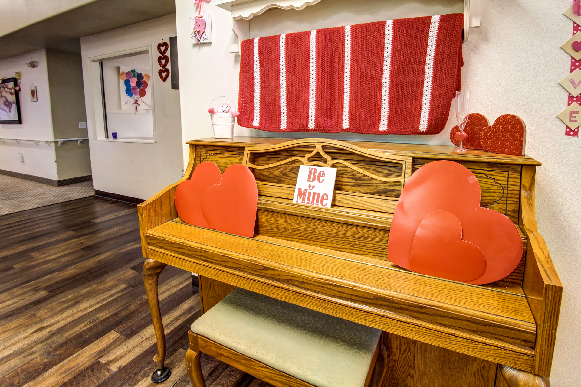 An oak piano decorated with red heart-shaped cutouts and a small sign that says 'Be Mine'. A red and white striped knitted blanket hangs on the wall above the piano. The setting appears to be a hallway with wood flooring and light-colored walls, decorated with Valentine's Day themed items.