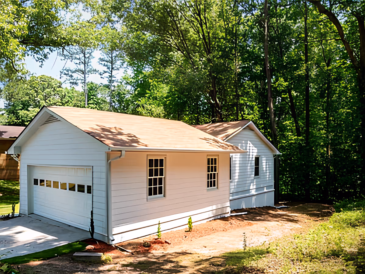 Exterior view of a white single-story house with a garage, surrounded by tall green trees and a dirt yard area.