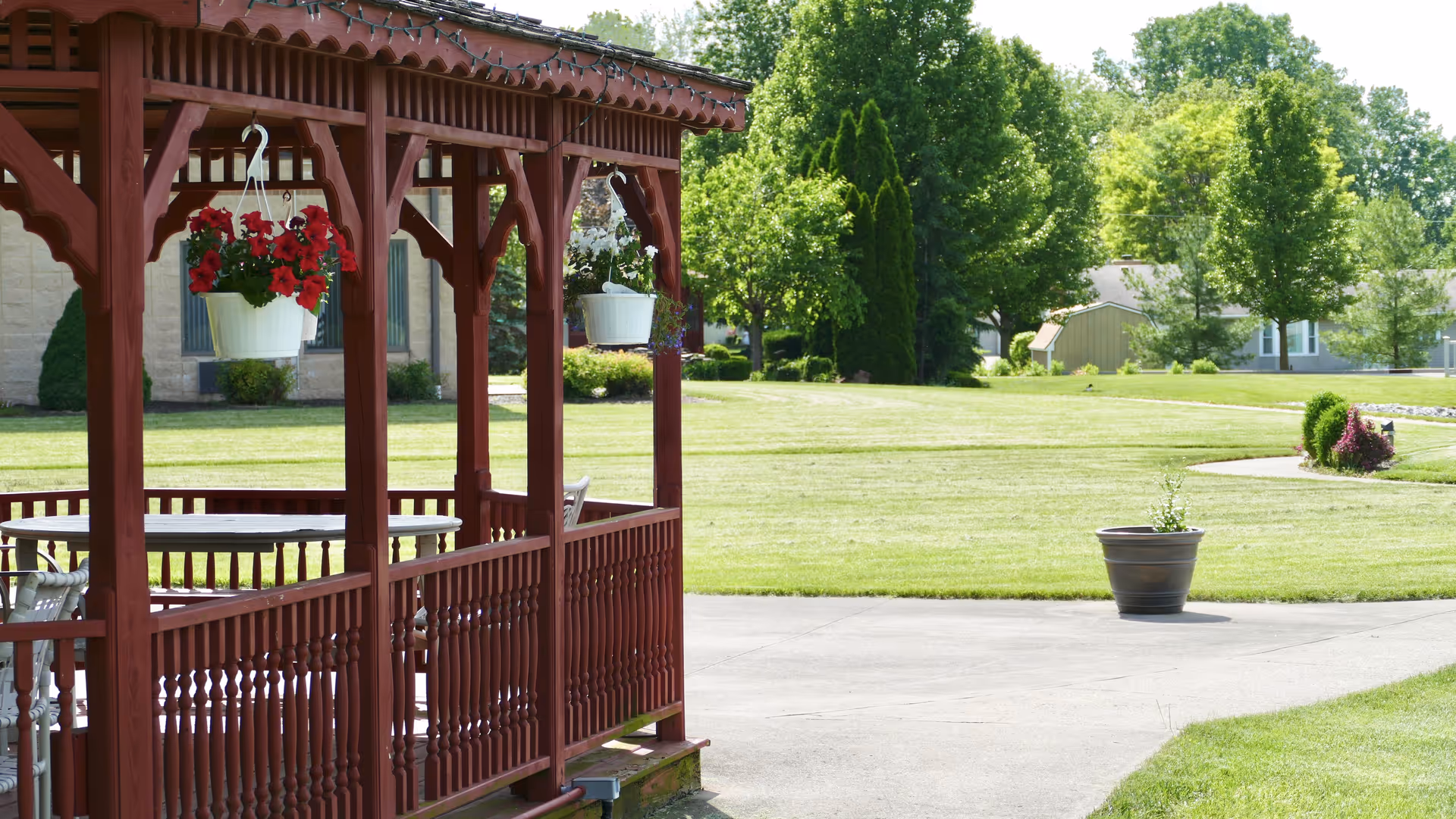 A wooden gazebo with hanging flower pots containing red and white flowers, situated on a concrete patio. In the background, there is a well-maintained grassy lawn with trees and residential buildings under a clear sky.