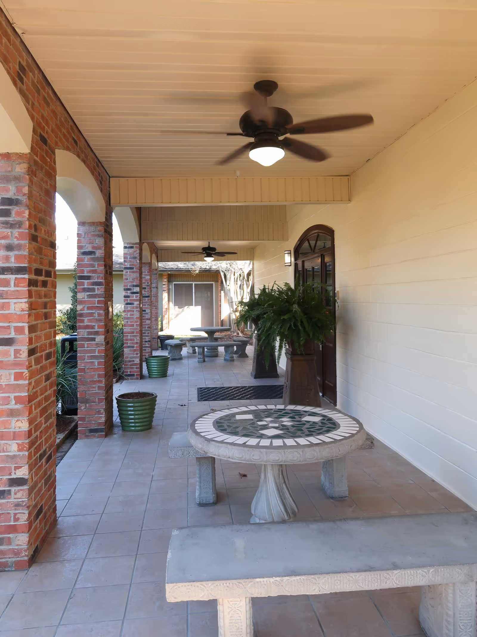Covered outdoor patio area with tiled floor, brick columns, ceiling fans, mosaic stone tables with matching benches, and potted plants along the wall and columns.