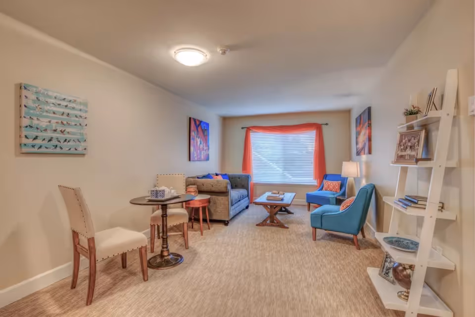 A cozy living room with beige walls and carpeted floor. The room features a small round table with two beige upholstered chairs on the left, a gray sofa with colorful cushions, a wooden coffee table, and two blue armchairs with orange patterned pillows. A large window with orange curtains lets in natural light. There are colorful paintings on the walls and a white ladder-style bookshelf holding books, framed photos, and decorative items on the right.