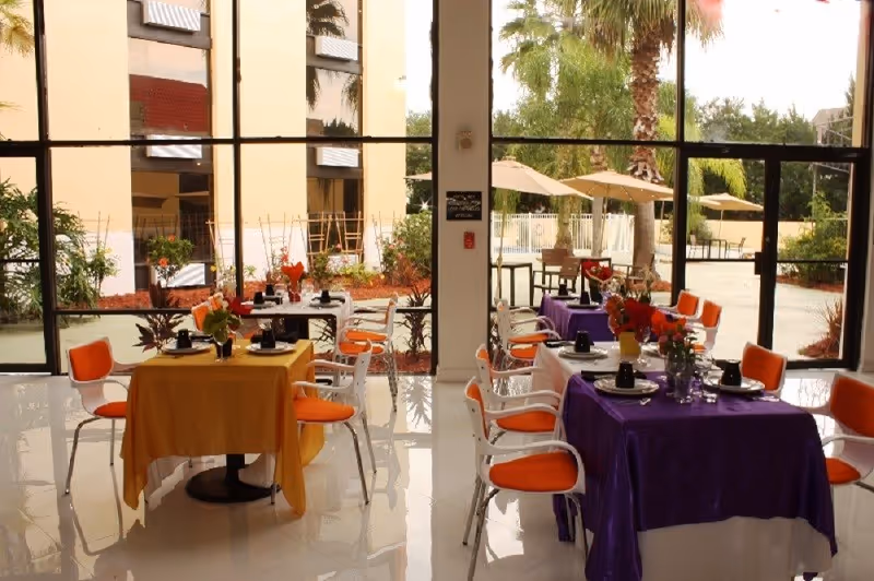 Bright dining area with orange and purple tablecloths, orange chairs, and large windows.