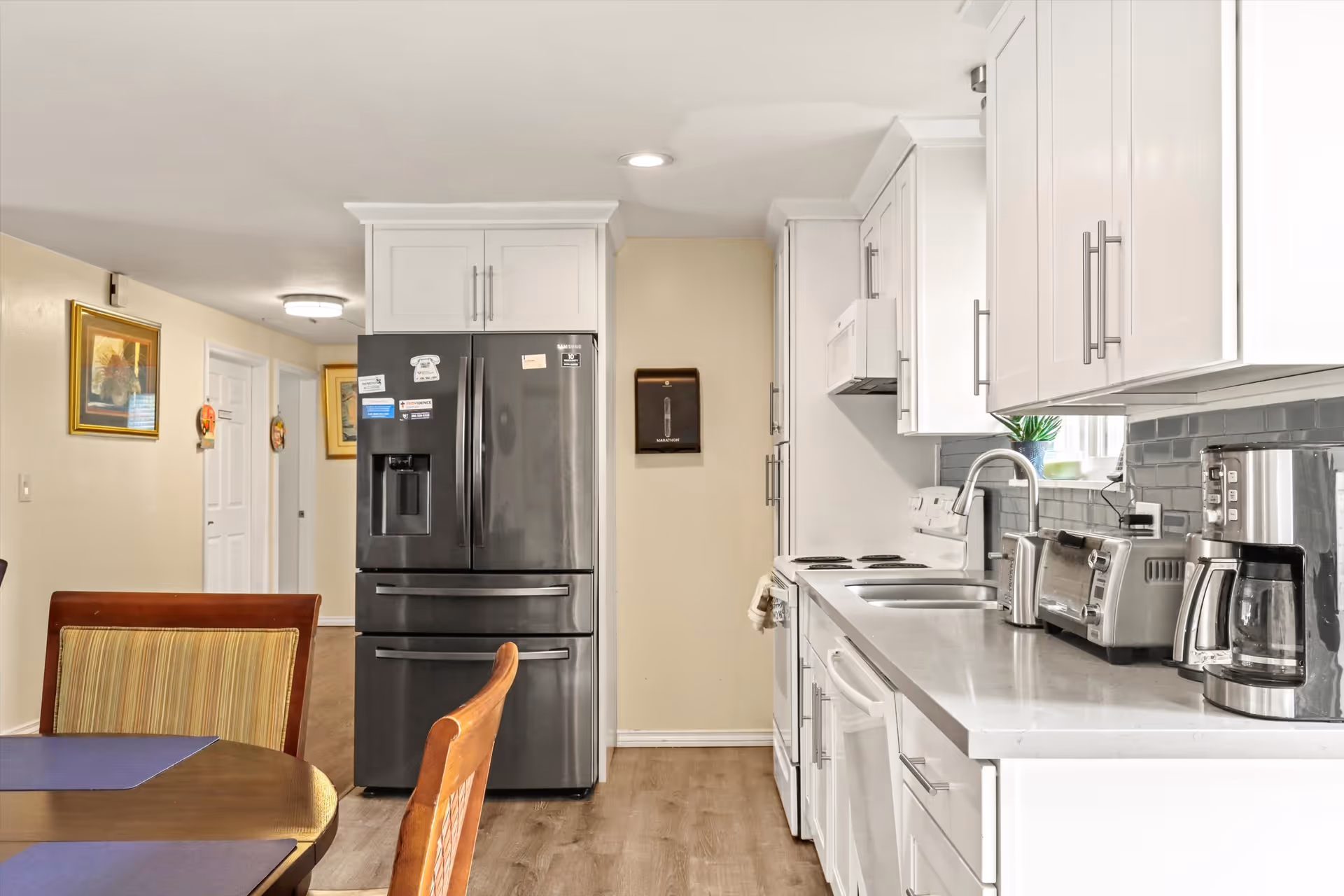Modern kitchen with white cabinets, a stainless steel refrigerator, a double sink, a coffee maker, toaster, and microwave. A wooden dining table with chairs is partially visible in the foreground. The kitchen has a light-colored backsplash and wood flooring.