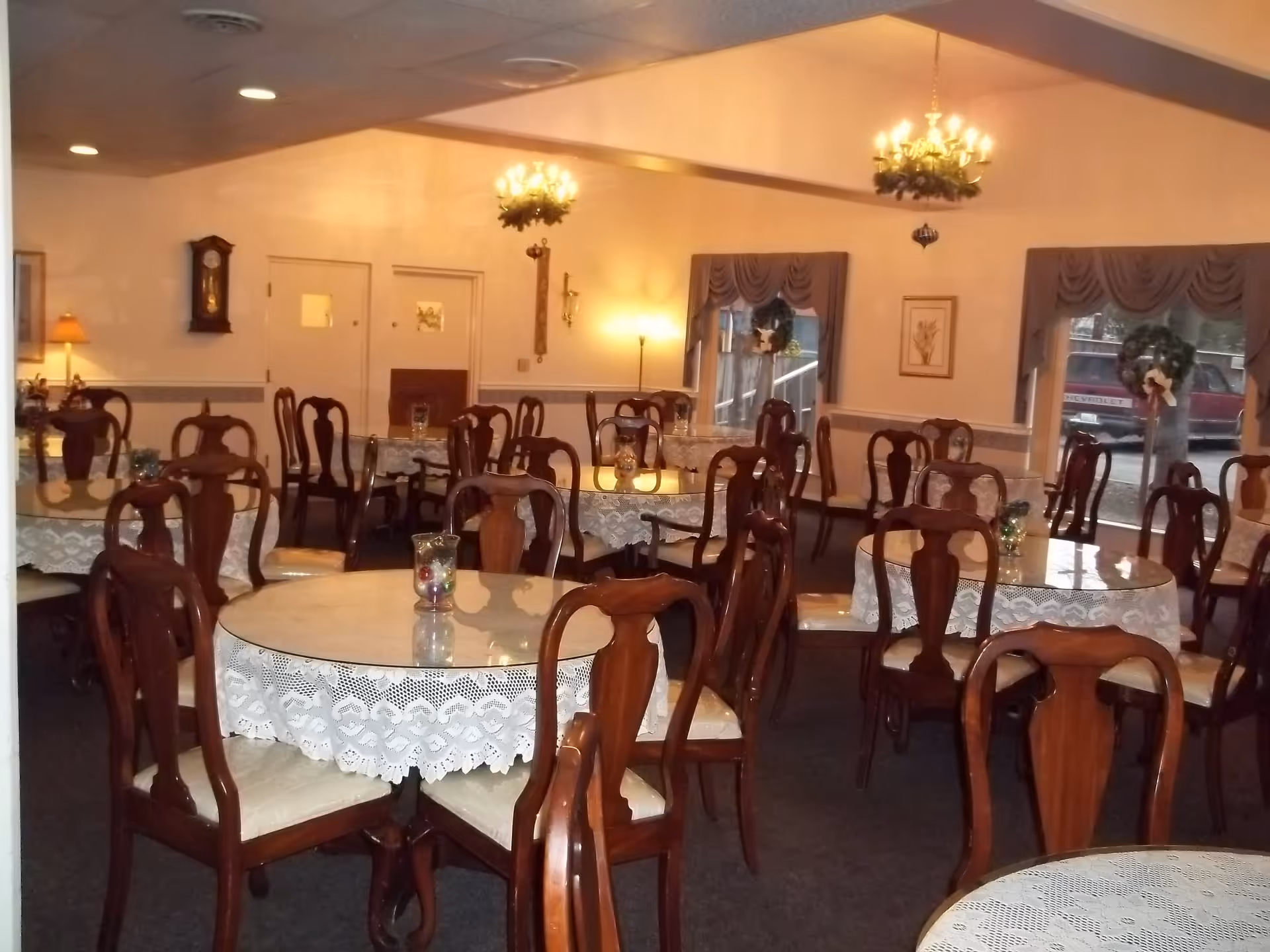 Interior view of a dining room with multiple round tables covered with white lace tablecloths and wooden chairs with cushioned seats. The room is warmly lit with chandeliers and wall lamps, and windows with draped curtains show a view outside. Decorative items like a grandfather clock and framed pictures are on the walls.