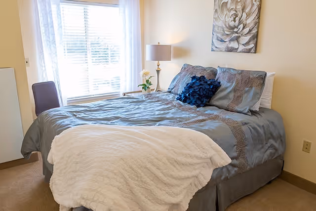 Neatly made bed with blue-gray bedding, decorative pillows, a bedside lamp and wall art next to a window with blinds and sheer curtains.