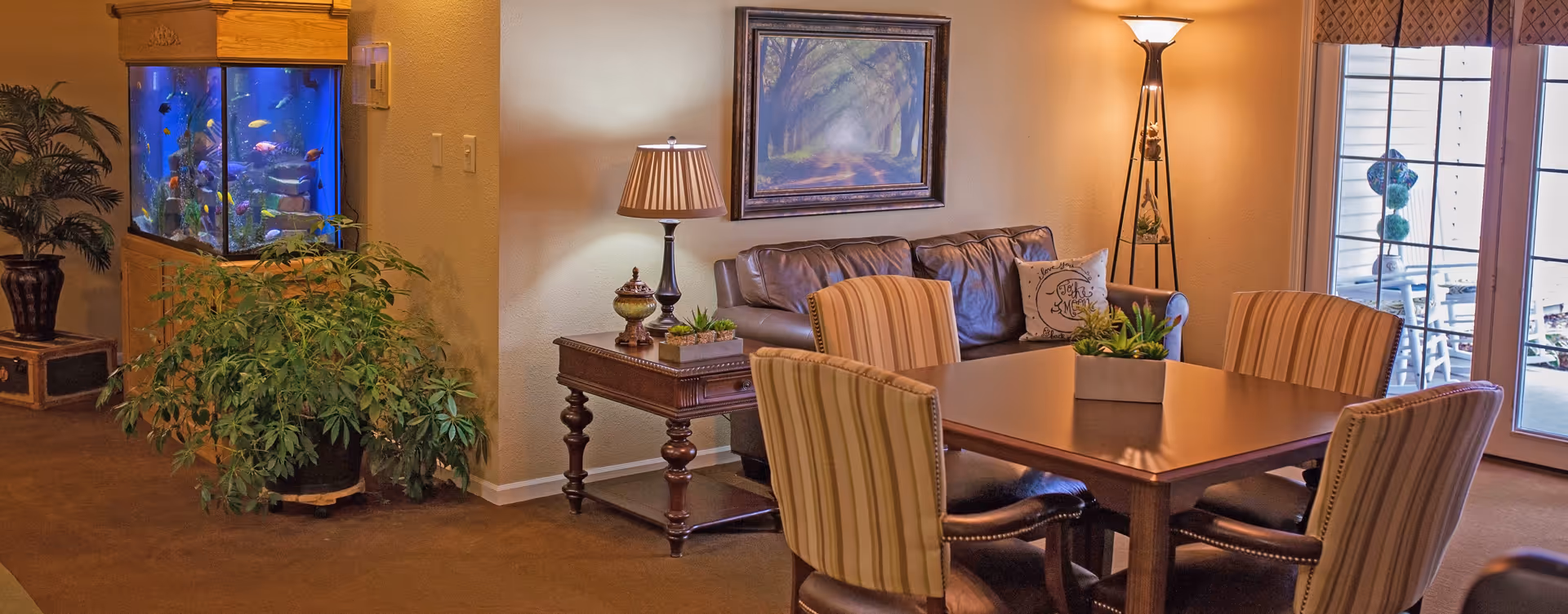 A cozy living room area with a brown leather sofa, a wooden side table with a lamp and decorative items, a wooden dining table surrounded by four striped upholstered chairs, a large fish tank built into the wall, several green plants, a floor lamp, and a framed painting of a tree-lined path on the wall. French doors with window blinds lead to an outdoor patio.
