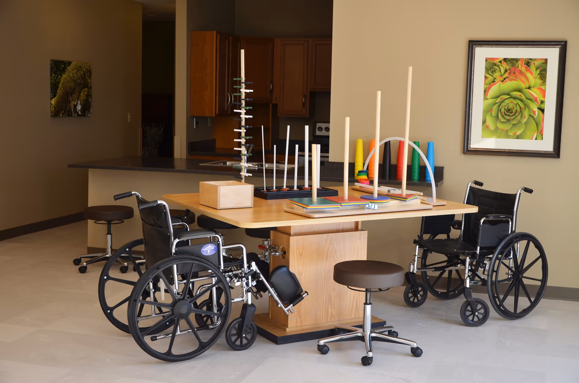 A therapy or activity room with a wooden table holding various occupational therapy tools and games. Two black wheelchairs and a brown rolling stool are positioned around the table. The background shows a kitchen area with wooden cabinets and a framed picture of a green succulent plant on the wall.