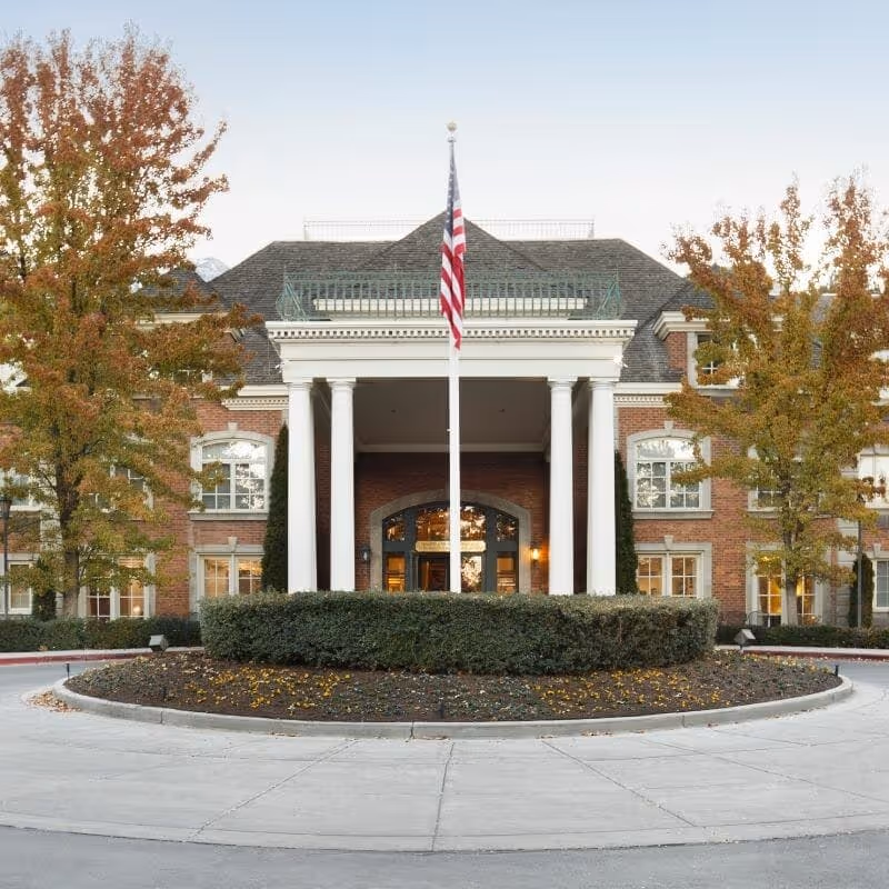 Front exterior view of a large brick building with white columns at the entrance, an American flag on a flagpole in the center, and trees with autumn foliage on either side.