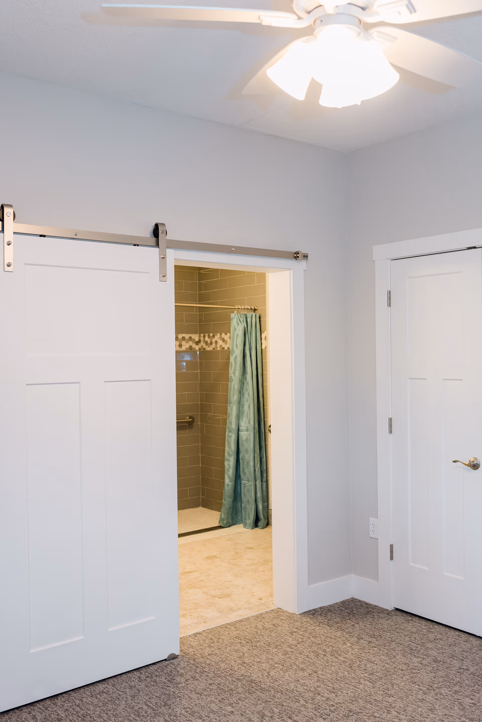 Carpeted interior room with a sliding barn door opened to a tiled bathroom showing a teal shower curtain and a ceiling fan above.