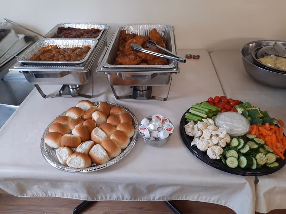 A buffet table with trays of fried chicken, meatballs, a platter of dinner rolls, a small bowl of creamers, and a vegetable tray with celery, cherry tomatoes, broccoli, cauliflower, carrots, and cucumber slices with a bowl of dip in the center.