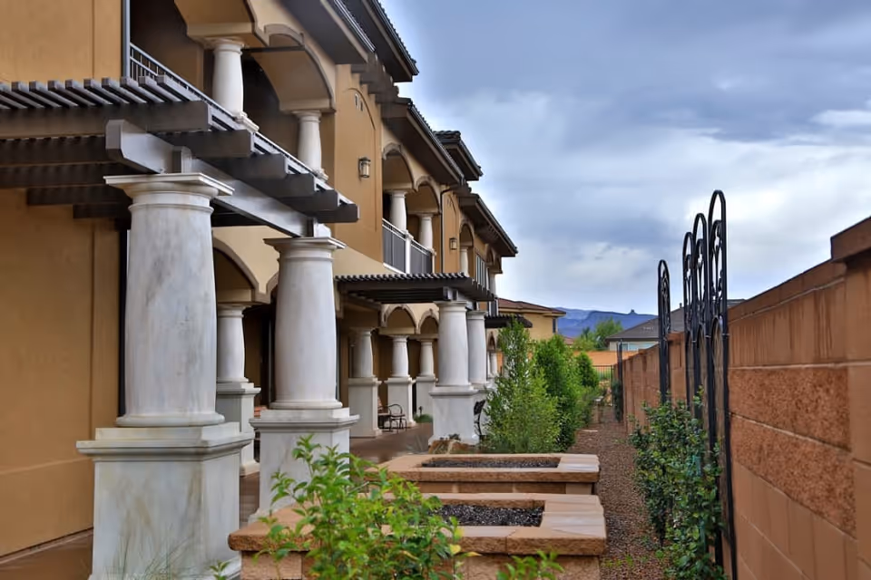 Exterior walkway and courtyard of a multi-story building with large white columns, raised planters, and a brick wall under a cloudy sky.