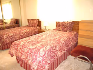 A bedroom with a single bed covered in a red and beige patterned bedspread and matching pillow. Next to the bed is a wooden nightstand with a lamp. There is a wooden dresser and a red cushioned chair with metal legs. A mirrored closet door is visible on the left side of the room.