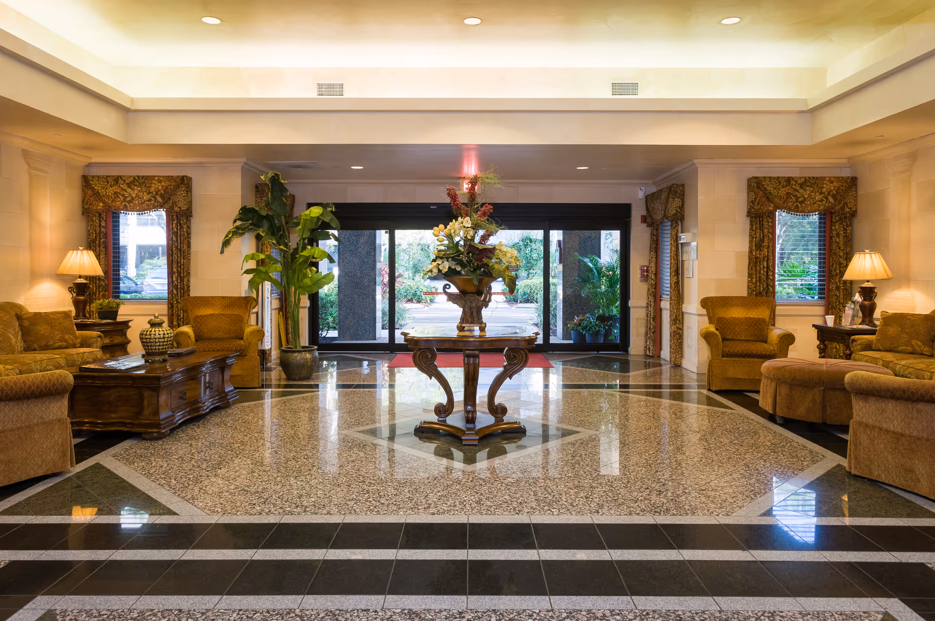 A spacious and elegant lobby area with polished tiled floors featuring a decorative pattern. In the center, there is a round wooden table with a large floral arrangement. The seating area includes upholstered armchairs and sofas in warm tones, with side tables and lamps. Large glass doors at the back open to an outdoor garden area, and there are windows with patterned curtains on either side of the room.