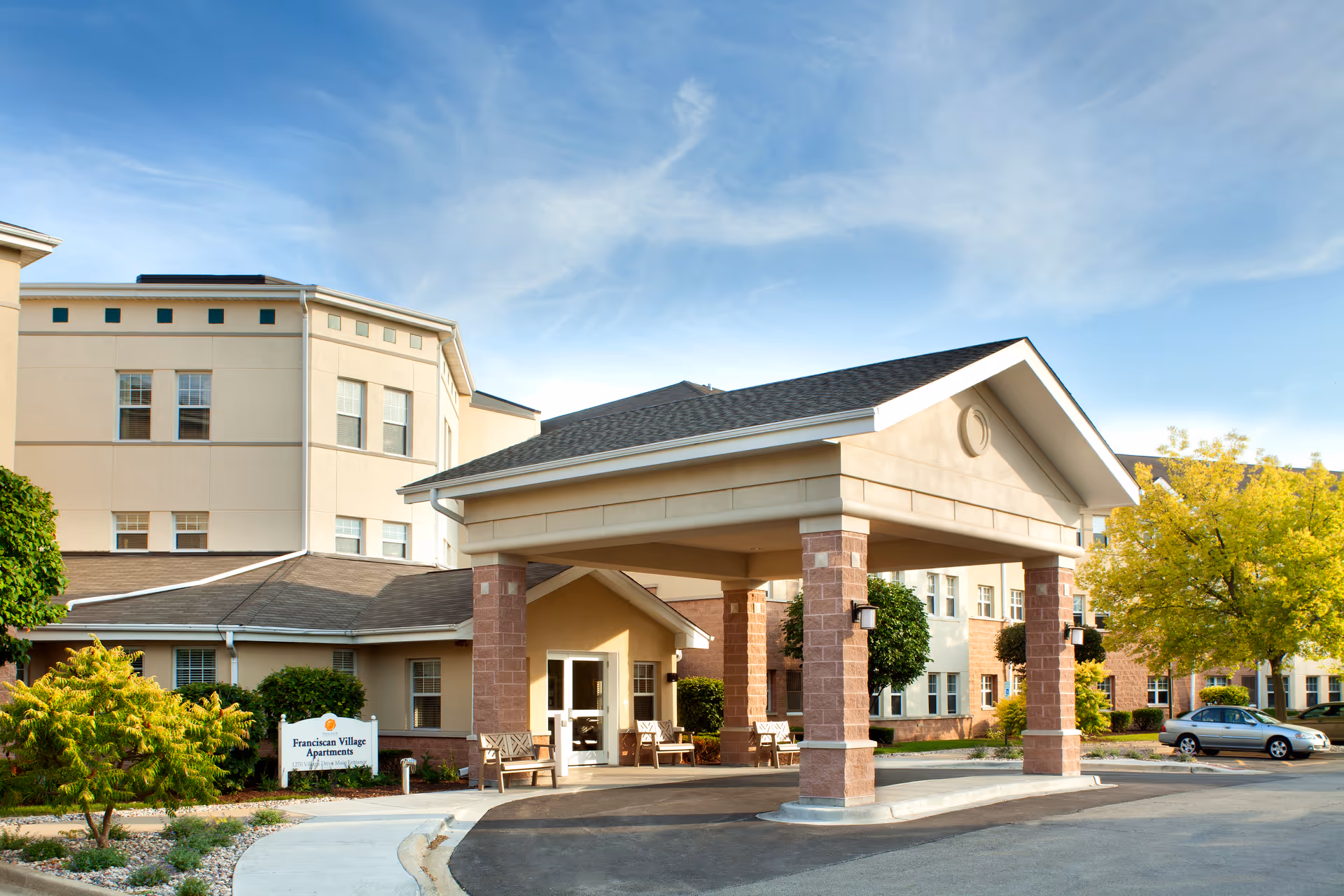 Exterior view of Franciscan Village Apartments showing the entrance with a covered driveway supported by brick pillars, benches near the entrance, surrounding greenery, and a parked car under a clear blue sky.