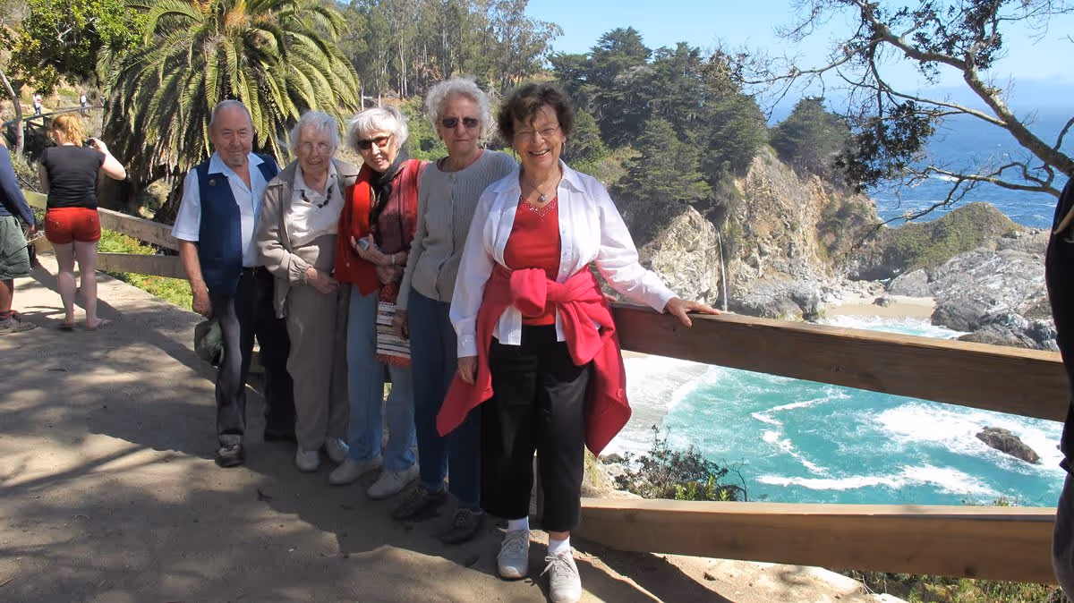 A group of five elderly people standing on a scenic outdoor trail with a wooden railing, overlooking a rocky coastline with turquoise ocean waves and trees in the background. Other people are visible in the background on the trail.