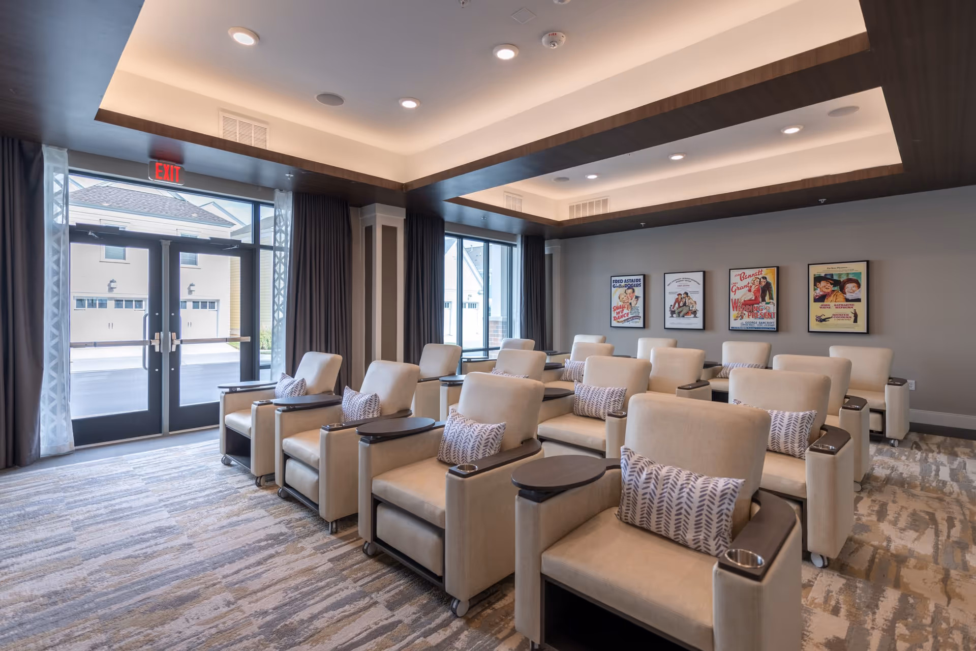 A cozy, modern common room with multiple beige armchairs arranged in rows, each with a patterned pillow and a small attached table with a cup holder. The room has large windows with dark curtains, a recessed ceiling with lighting, and framed vintage movie posters on the wall. Double glass doors lead outside to a paved area with garages visible.