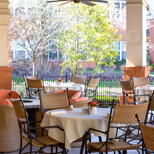 Outdoor covered patio area with several round tables covered with white tablecloths, surrounded by wicker and metal chairs. Each table has a small flower arrangement and rolled napkins. The patio overlooks a garden with trees and shrubs, and a building with windows is visible in the background.