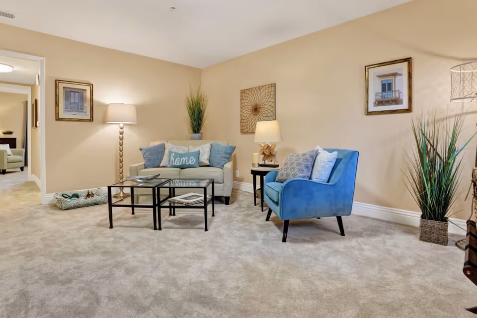 A cozy living room with beige walls and carpeted floor featuring a small sofa with decorative pillows, including one with the word 'home', a blue armchair with patterned cushions, two glass-top nested coffee tables, a floor lamp, a side table with a lamp and decorative items, framed artwork on the walls, and tall green plants in woven baskets.