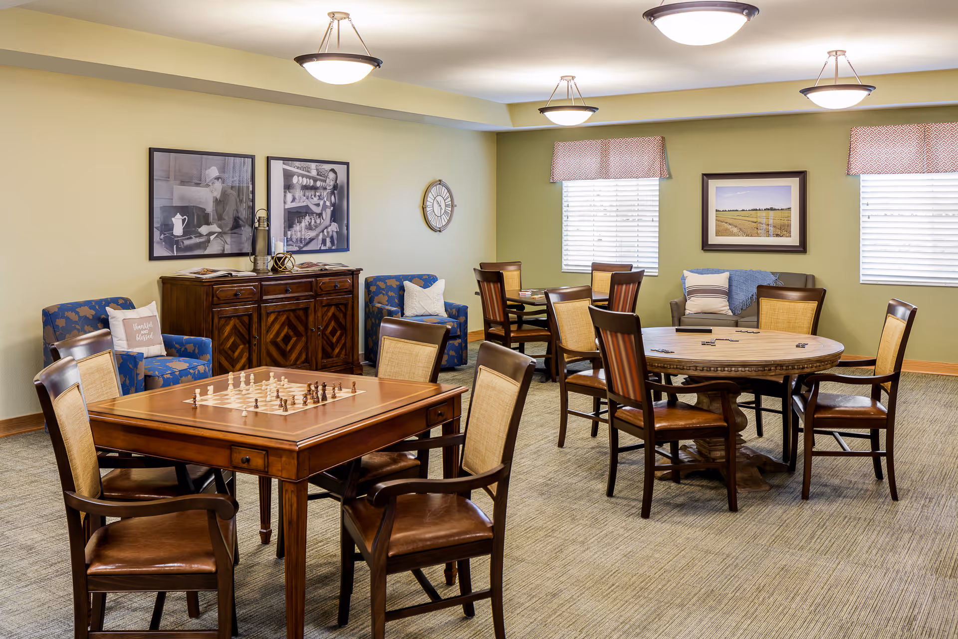 A cozy common room with two wooden tables surrounded by chairs, one table set up for chess and the other with dominoes. The room has two blue patterned armchairs with pillows, a wooden cabinet with decorative items, framed black and white photos on the wall, a clock, and two windows with blinds and patterned valances. The walls are painted light green and beige, and the room is well-lit with ceiling lights.
