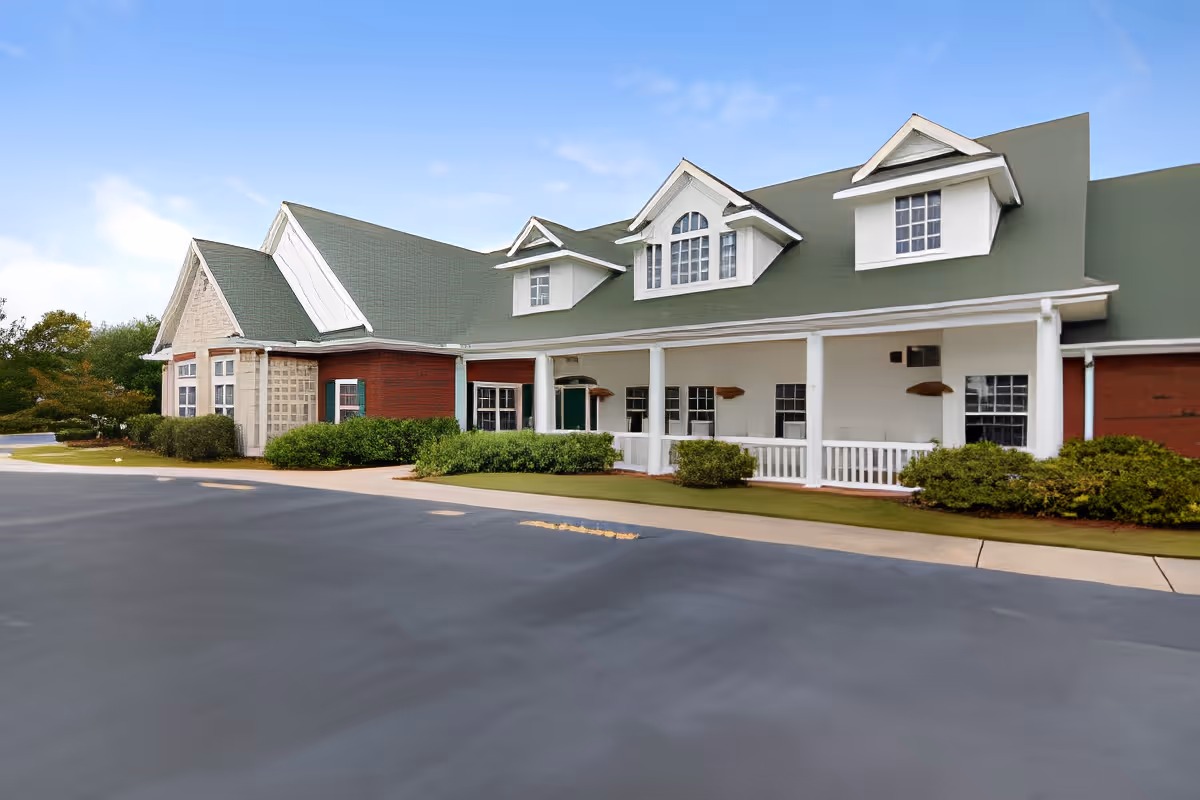 Exterior view of a single-story building with a green roof, white columns, and red brick walls, surrounded by bushes and a paved driveway under a blue sky.