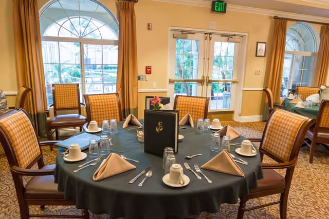 A dining room with a round table set for six people. The table is covered with a dark green tablecloth and has beige napkins folded in a triangle shape, white cups and saucers, glasses, and silverware arranged neatly. The room has large arched windows with yellow curtains, patterned carpet, and several wooden chairs with cushioned seats and backs. There is a menu or decorative item in the center of the table and an exit door in the background.