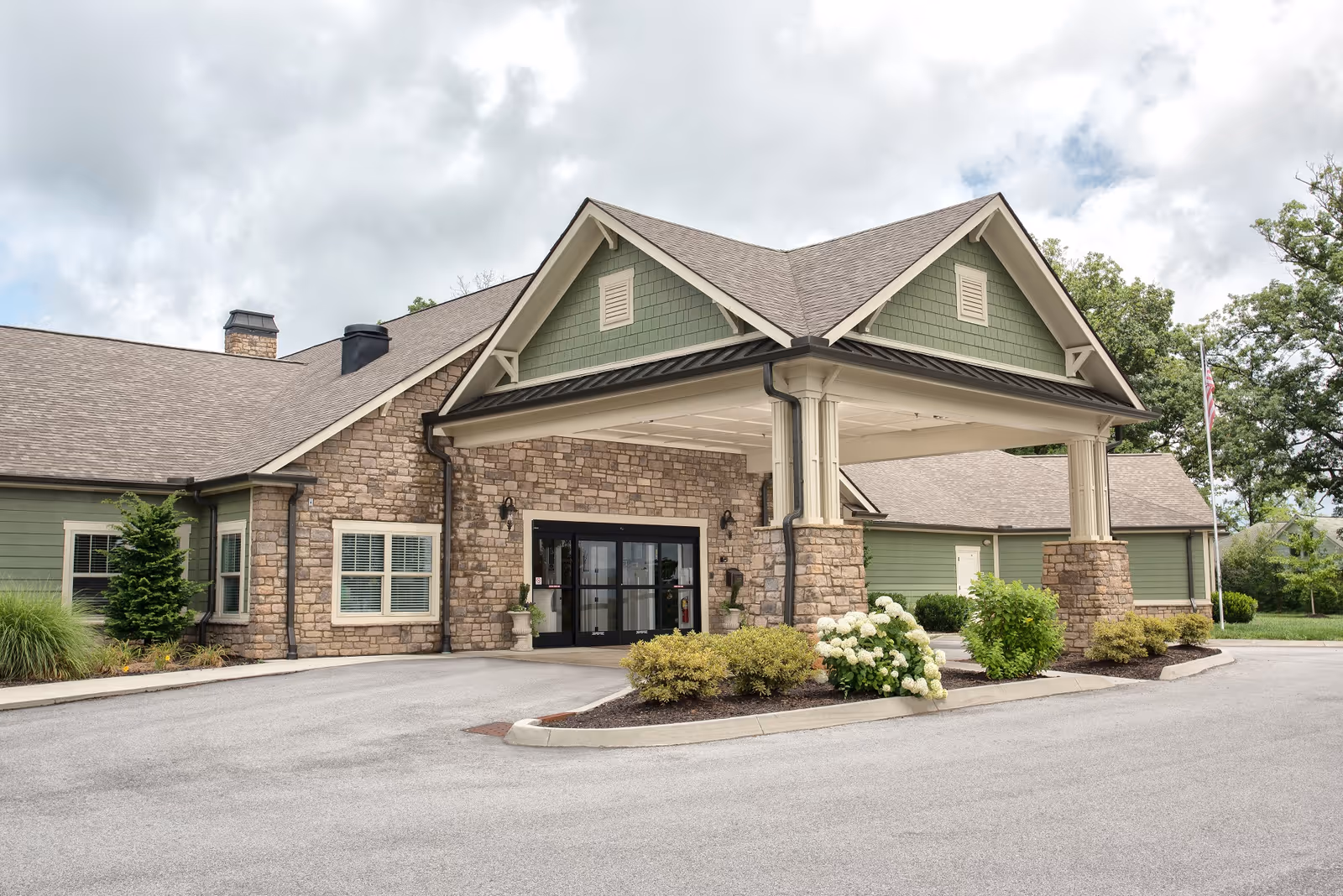 Exterior view of Clover Hill Senior Living facility showing a stone and green siding building with a covered entrance, landscaped bushes, and an American flag on a flagpole under a cloudy sky.