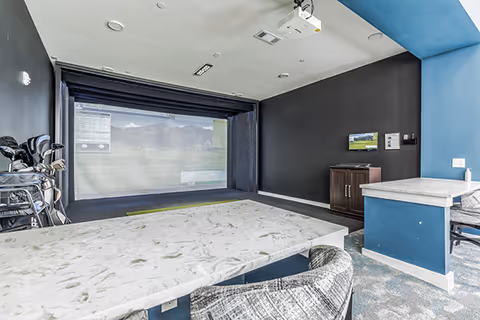 Indoor golf simulator room with a large screen displaying a golf course, golf clubs on a rack to the left, a marble table with patterned chairs in the foreground, and a blue accent wall with a small cabinet and a mounted screen on the right.