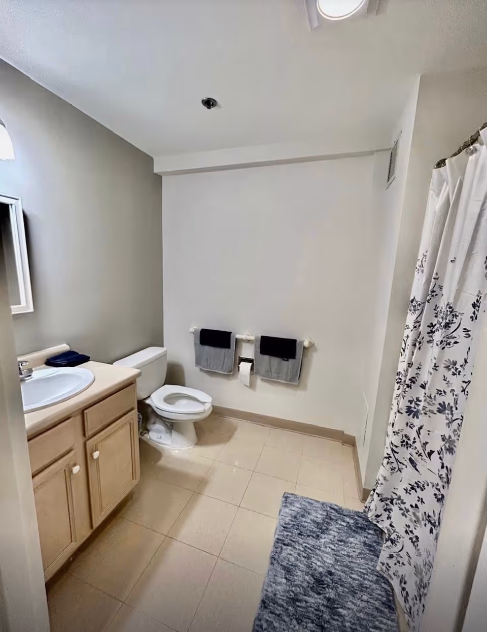 A clean bathroom with beige tiled floor, a white toilet, a wooden vanity with a sink and mirror, two towel racks with gray and black towels, a shower with a white curtain featuring a blue floral pattern, and a blue bath mat on the floor.