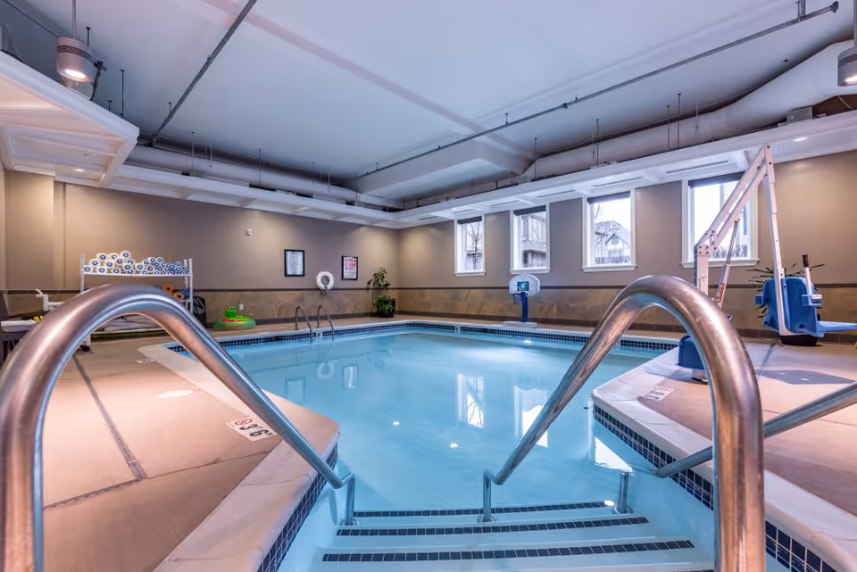 Indoor swimming pool with metal handrails leading into the water, surrounded by beige tiled flooring and walls. The pool area has several windows letting in natural light, a pool lift for accessibility, and pool equipment stored on a rack in the corner.
