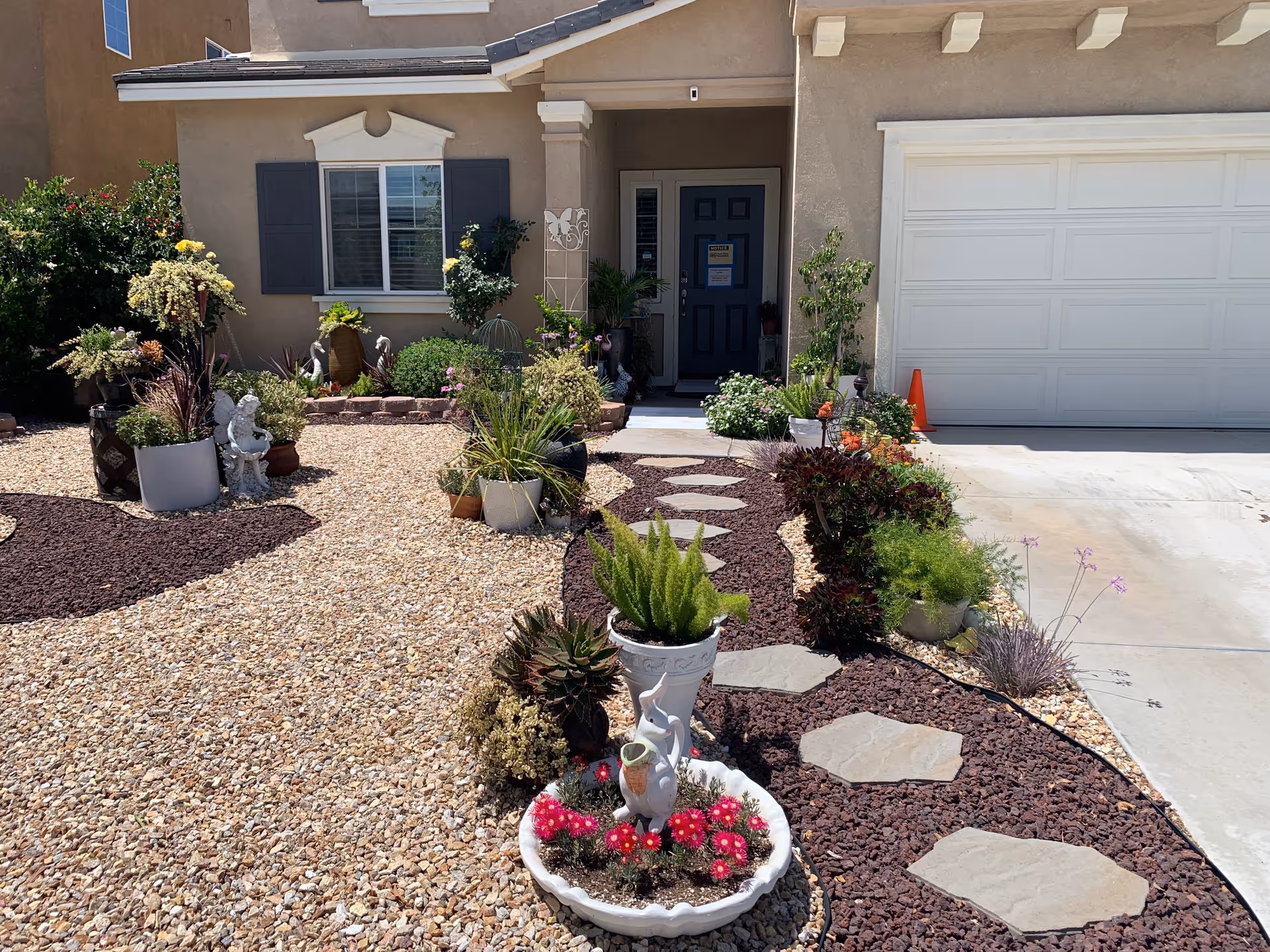 Front exterior view of a beige house with a dark blue door, white garage door, and a landscaped front yard featuring various potted plants, succulents, flowers, and a stone pathway leading to the entrance.