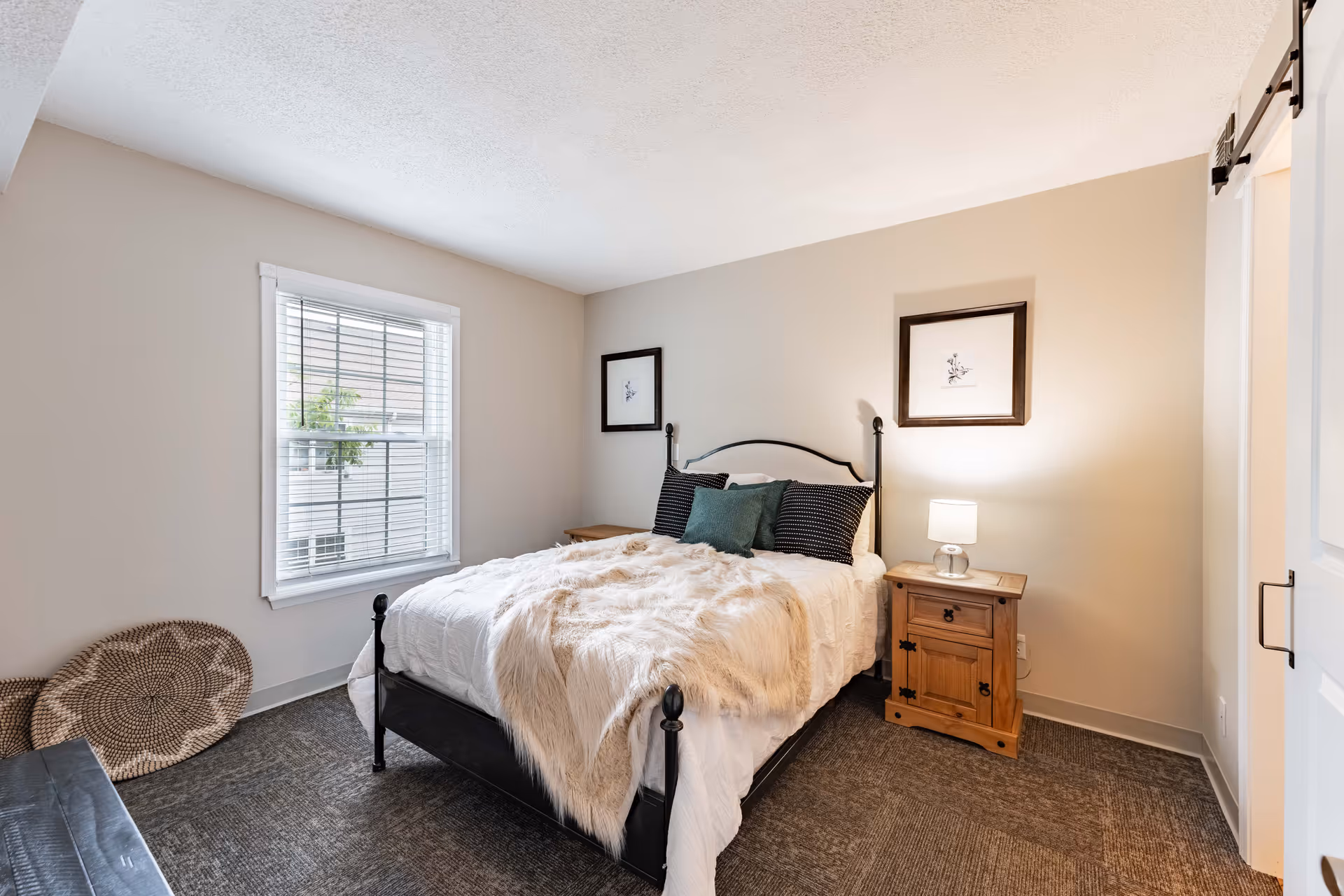Cozy bedroom with an iron-frame bed draped in a fur throw, a wooden nightstand with a lamp, a window with blinds, and framed artwork on the wall.